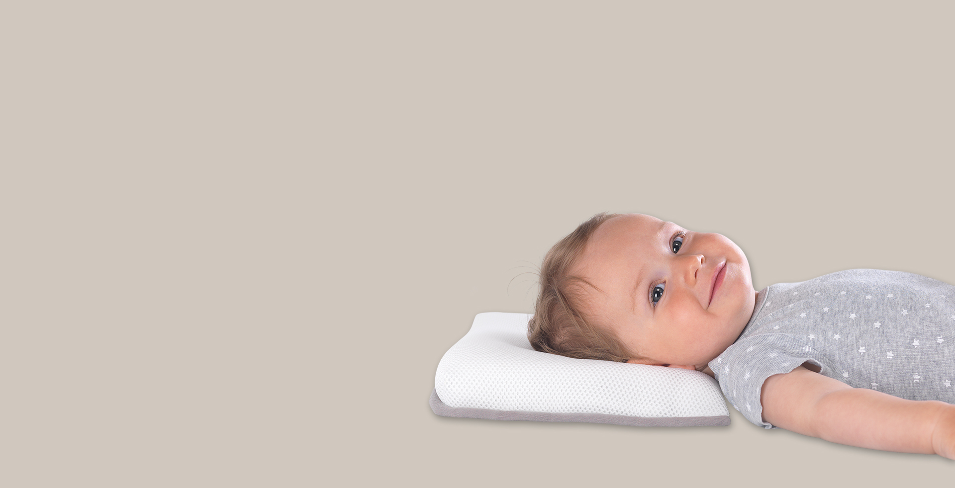 Ein Baby liegt lächelnd auf einem weißen Theraline-Kopfkissen, gekleidet in ein graues Oberteil mit kleinen Sternenmustern, vor einem beigefarbenen Hintergrund.
