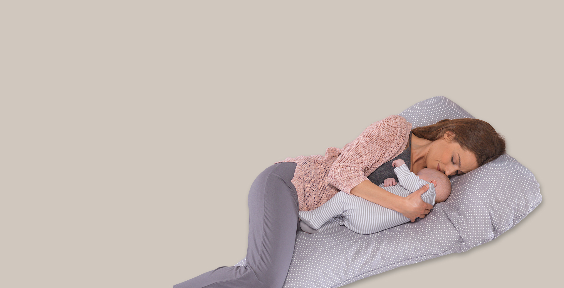 Eine Frau liegt schlafend mit einem Baby im Arm auf einem Theraline-Kissen mit grauem, gepunktetem Bezug. Der Hintergrund ist einfarbig beige.