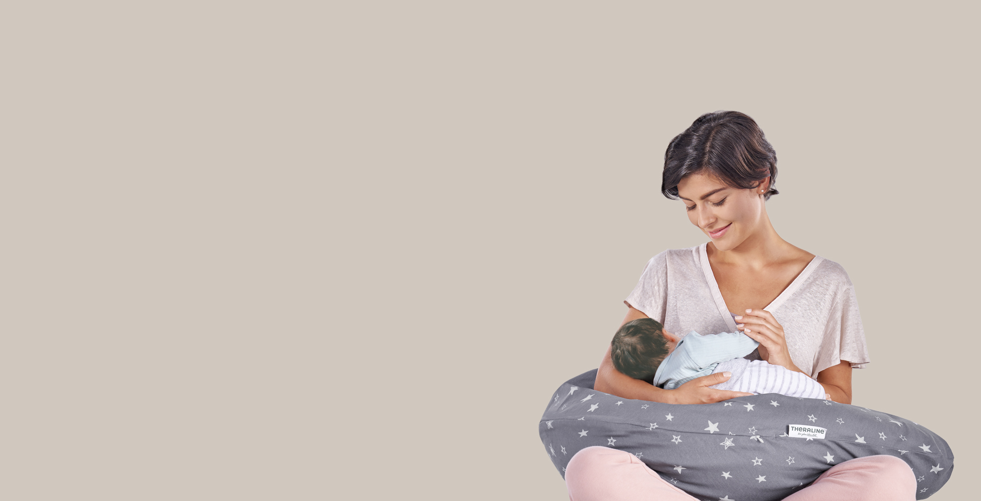 Eine Frau hält ein Baby auf einem grauen Stillkissen mit Sternenmuster. Sie sitzt lächelnd in einem neutralen Raum. Auf dem Kissen ist ein Etikett mit dem Markennamen 