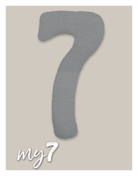 Ein graues, kissenartiges Objekt in Form einer Zahl 7 liegt auf einem beigen Hintergrund. Unten links befindet sich der Text 