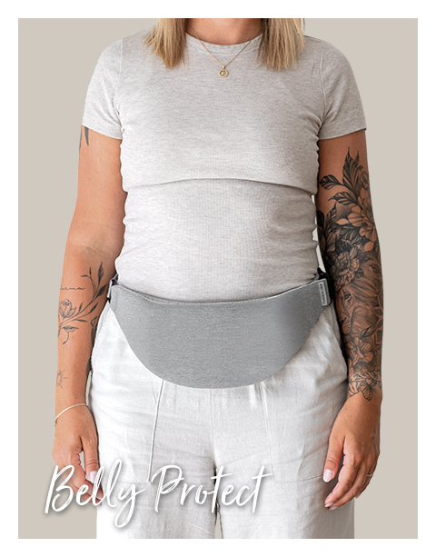 Eine Frau trägt eine Bauchschutzhülle von Theraline in Grau über ihrem T-Shirt, mit tätowierten Armen und einem Hintergrund in neutralem Ton. Text: