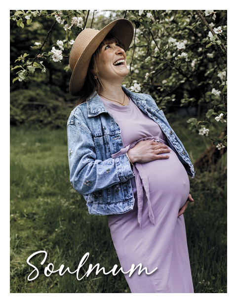 Eine schwangere Frau in einem lila Kleid und einer Jeansjacke hält ihren Bauch und lacht fröhlich. Sie steht in einem blühenden Garten. Text: